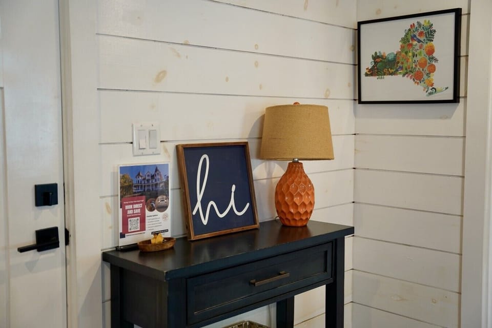 A warm entryway welcome to the barn unit, once a horse stable, with colorful New York state art and a cozy honeycomb lamp on a rustic console table. Shiplap walls and thoughtful details set the tone for your relaxing stay at Parkway House Goshen.