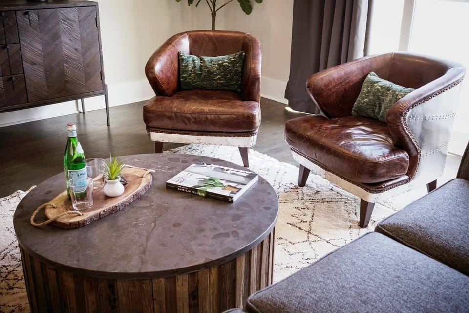 Sink into vintage charm with these cozy leather accent chairs, perfect for morning coffee or evening chats - Textured round coffee table brings earthy style and space to gather around - Styled with books, bubbly, & greenery for a warm, inviting touch