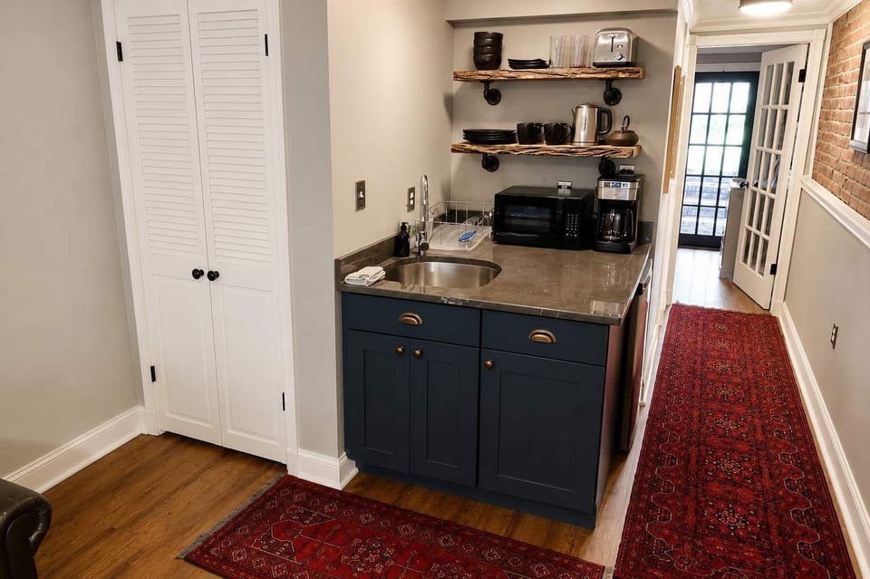 A charming, well-equipped kitchenette with a compact refrigerator, coffee maker, sink, and open shelving stocked with essential dishes and cookware. The warm wood floors and rich blue cabinetry add to the apartment’s cozy character.