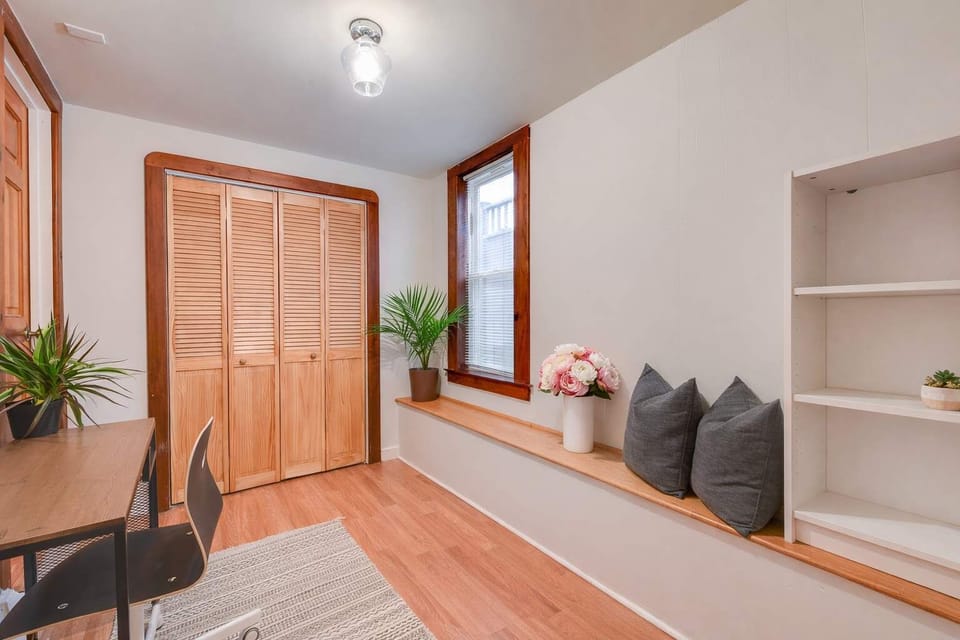 A bright dining nook with wood floors, built-in shelving, bench seating with cushions, leafy plants, and warm wood-trimmed windows.
