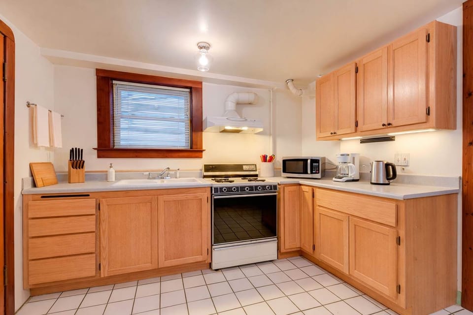 A bright, fully equipped kitchen with wood cabinetry, tiled floors, full-size stove, microwave, kettle, and ample counter space beneath a sunlit window.

