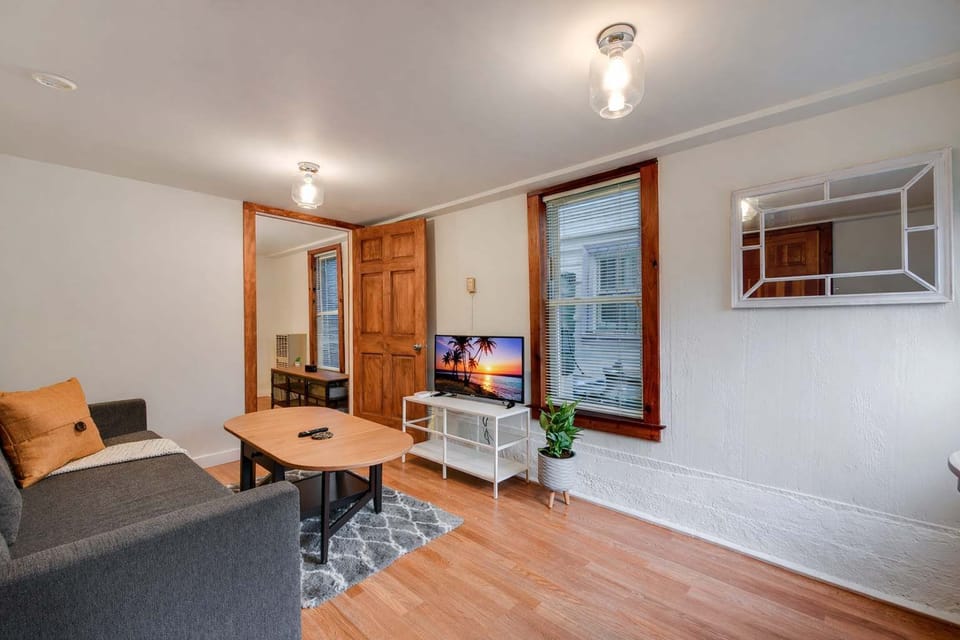 A bright, cozy living room with a gray sofa, patterned rug, coffee table, and TV stand, accented by warm wood trim, hardwood floors, and soft ceiling lighting.
