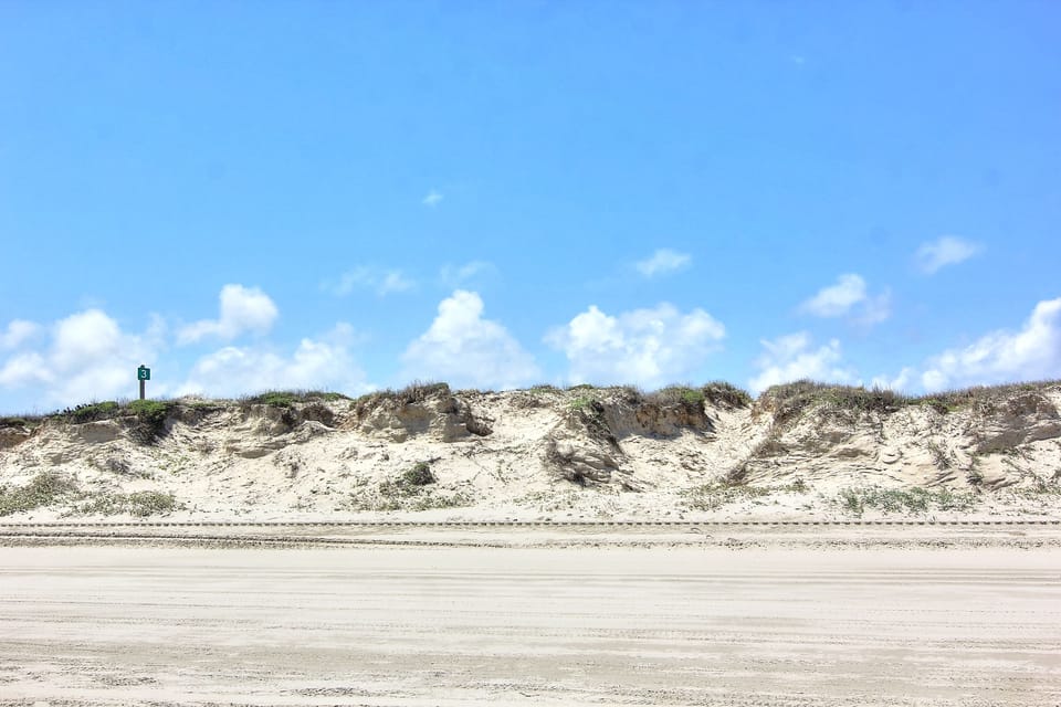 Natural coastal dunes rise above pristine sandy shoreline, creating a peaceful beachfront setting with clear blue skies.