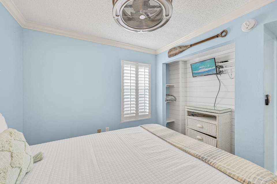 Your coastal-themed guest bedroom awaits with soothing blue walls and crisp white linens. The ceiling fan keeps you cool while you watch TV from your comfortable bed.