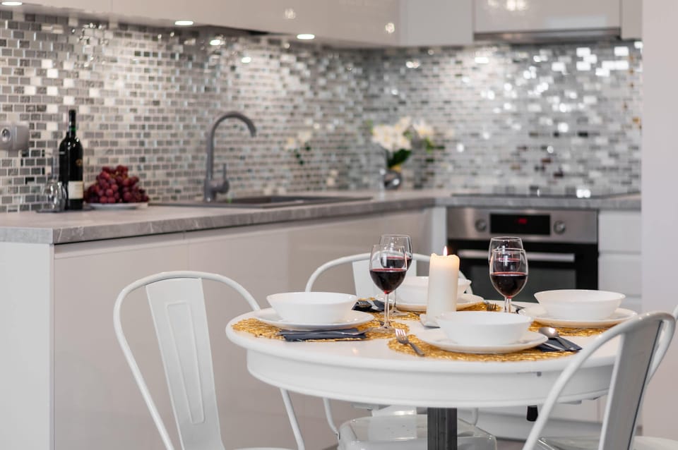 A stylish dining area adjacent to a modern kitchen with a small round table set for two, featuring white furniture and a sparkling chandelier overhead.
