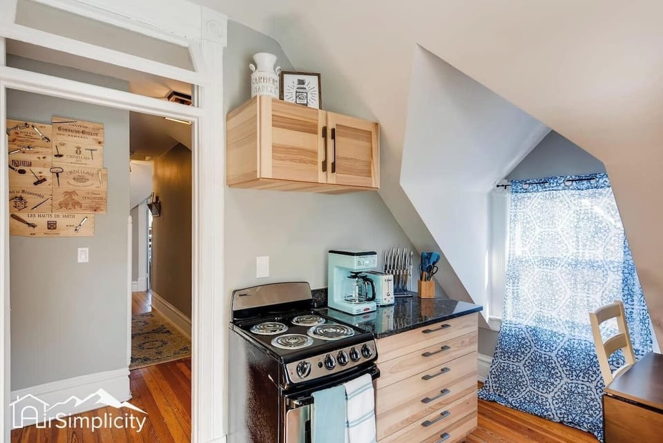 - Corner kitchenette includes a compact electric stove, prep space, and full utensil set
- Natural light from the window enhances the warmth of the soft wood cabinetry
- Coffee maker and toaster sit ready for quick breakfasts or late night snacks