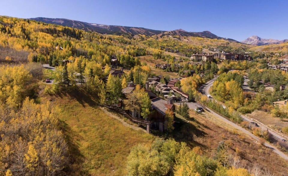 Aerial view of a residential area surrounded by autumn foliage and mountains in the background. Several houses are visible along winding roads amidst a landscape of yellow and green trees.