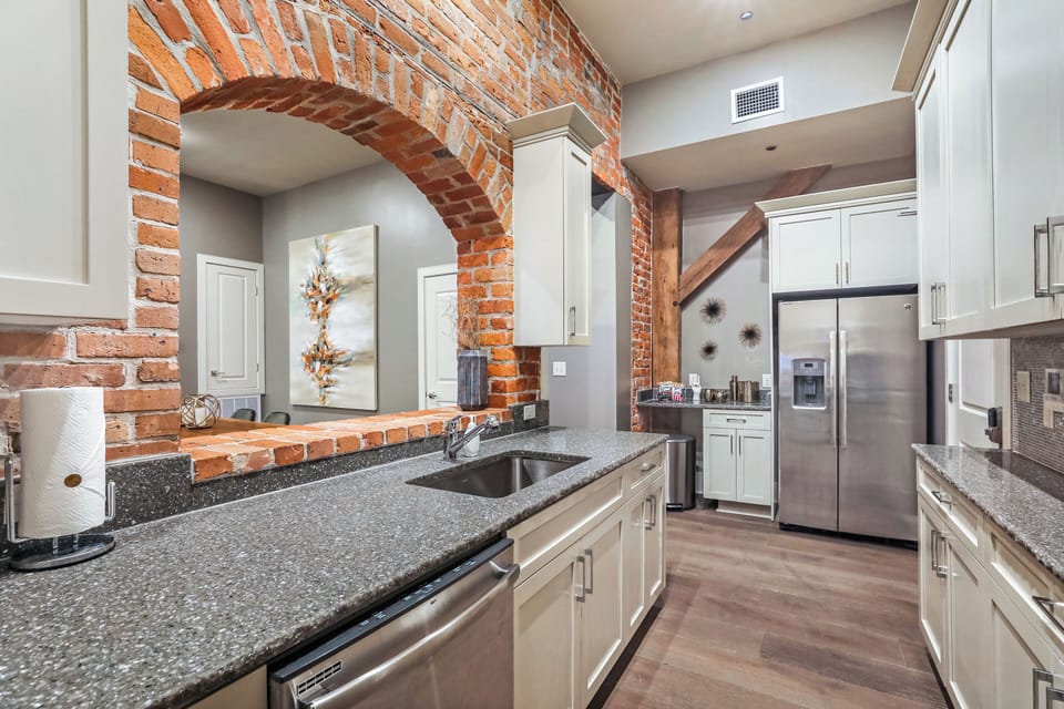 Galley-style kitchen open to living/dining area. Fully stocked with essentials from mixing bowls to wine glasses. Features stainless steel appliances, gas range, stone countertops, and tile backsplash, combining functionality with luxury.