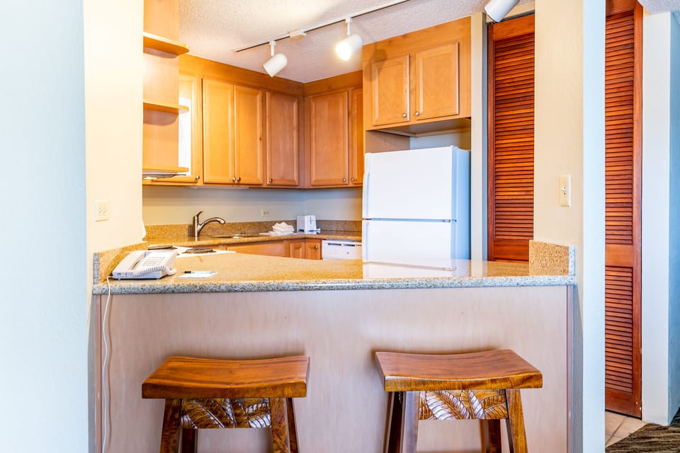 Your fully equipped kitchen features warm wood cabinetry, granite countertops, and modern appliances ready for meal preparation. A breakfast bar with handcrafted wooden stools creates the perfect spot for morning coffee or casual dining.
