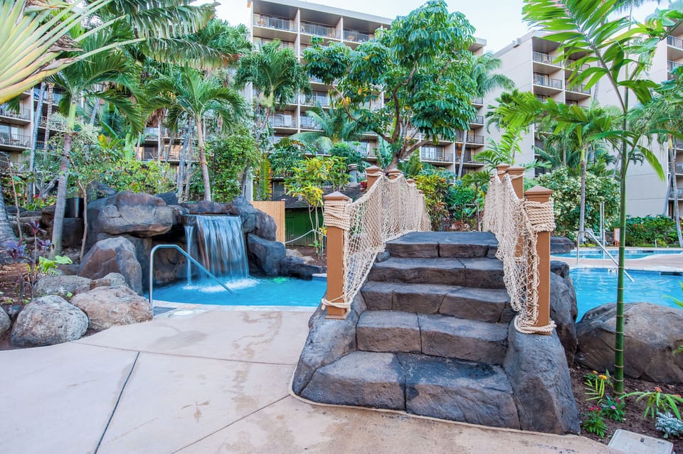 A tropical-themed pool area features a waterfall, rope bridge, and lush landscaping surrounded by multi-story resort buildings with balconies.