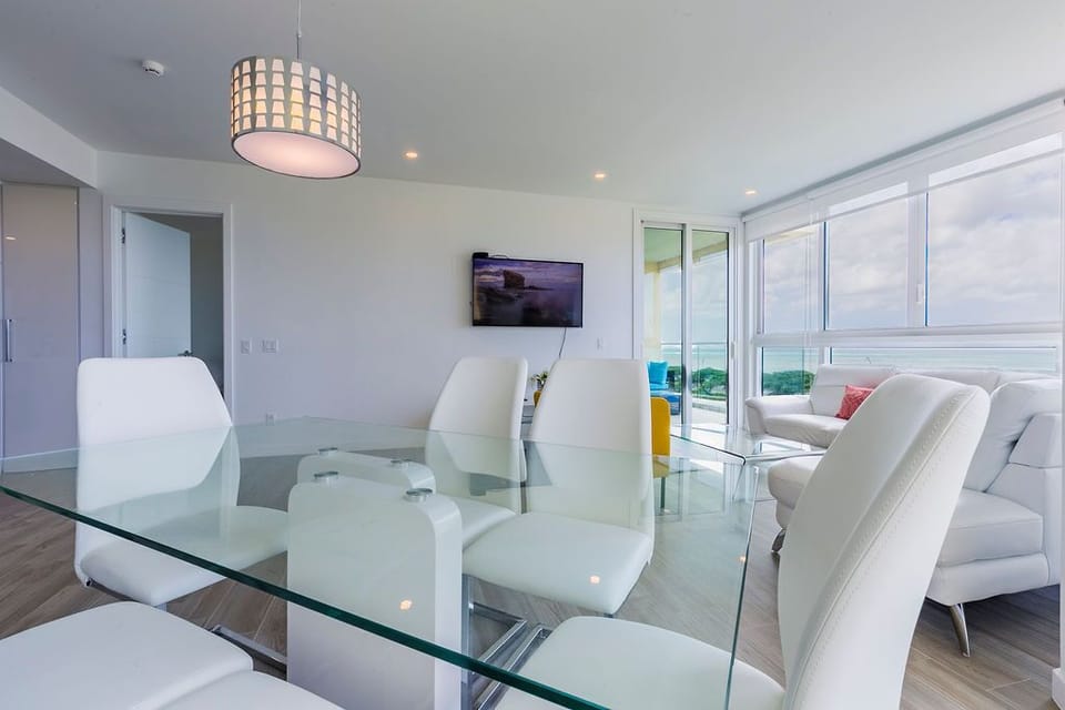 Your modern dining space features sleek white chairs and a glass table under soft pendant lighting. Floor-to-ceiling windows frame ocean views, while the open layout flows seamlessly into the living area.