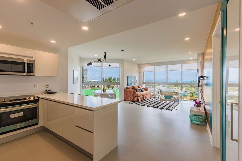 Modern open-concept space with sleek kitchen flowing into a living area, framed by floor-to-ceiling windows showcasing ocean views and tropical greenery beyond.