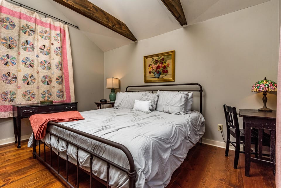 Bedroom with dark metal bed frame, white bedding, hardwood floors, vintage quilt hanging, and wooden desk with stained glass lamp.