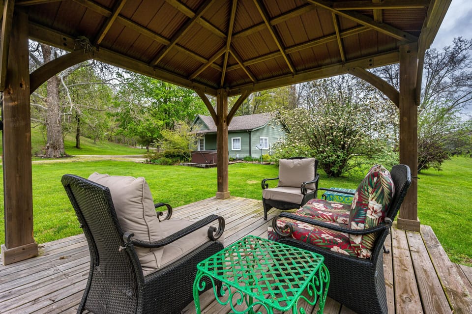 Wicker patio furniture with beige and floral cushions surrounds a green metal table under a wooden gazebo.
