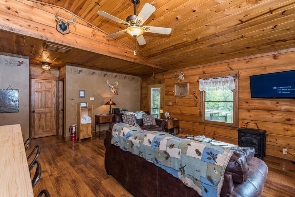 Wood-paneled living area with leather sofas, mounted flat-screen TV, ceiling fan, and windows overlooking greenery.