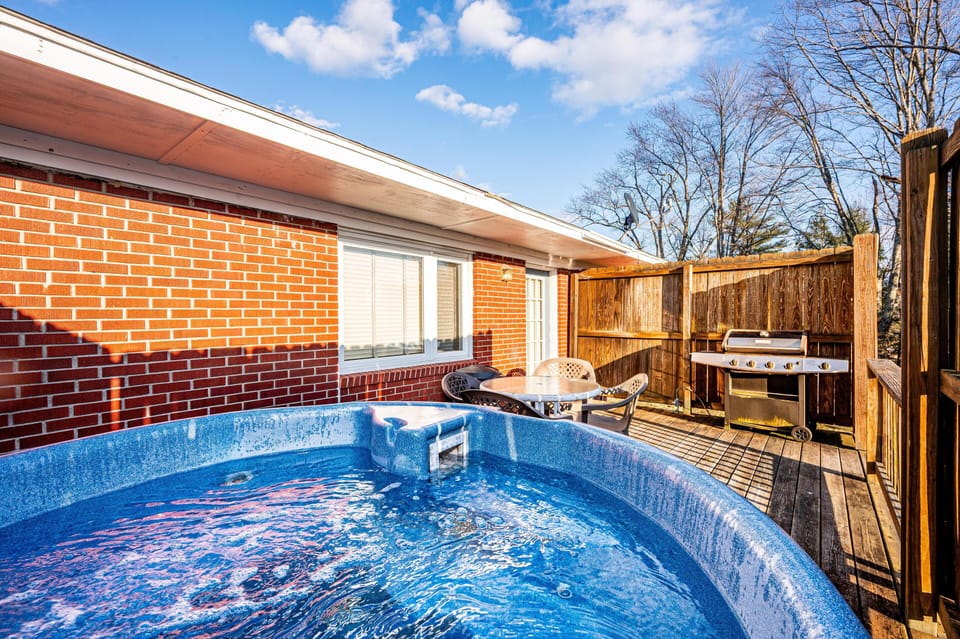 Outdoor deck with bubbling blue hot tub, round patio table with chairs, and stainless steel grill.