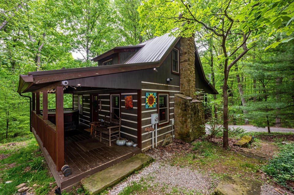Dark wood cabin exterior with stone chimney and covered porch featuring rustic bench and colorful wall art.