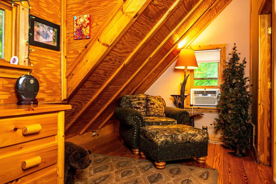 An armchair with matching ottoman, forest-themed upholstery, and wooden legs sits beneath a sloped pine ceiling.