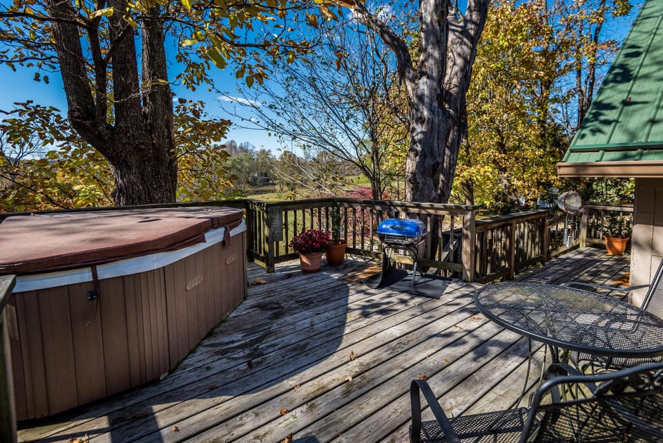 Wooden deck with round metal table and chairs, blue grill, potted plants, and covered hot tub under trees.