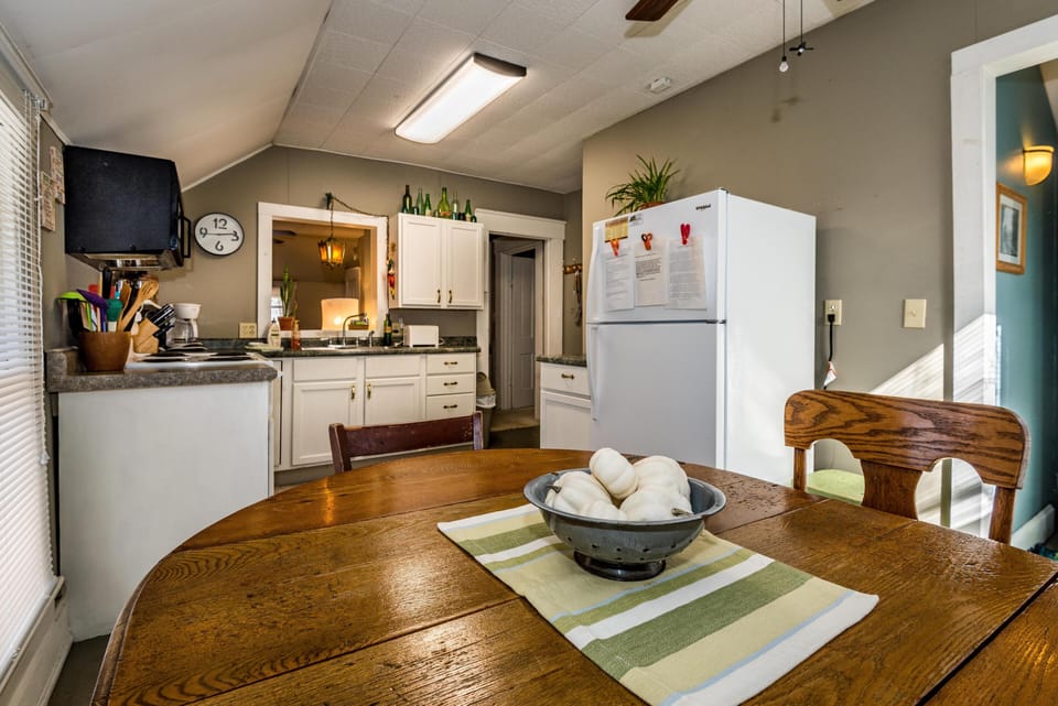 Wooden dining table topped with striped runner and white pumpkins, adjacent to white-paneled kitchen cabinets.