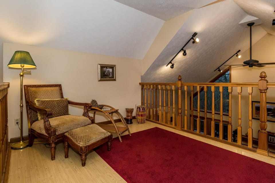 Upstairs nook with patterned armchair, matching ottoman, brass floor lamp, and red area rug by wooden railing.
