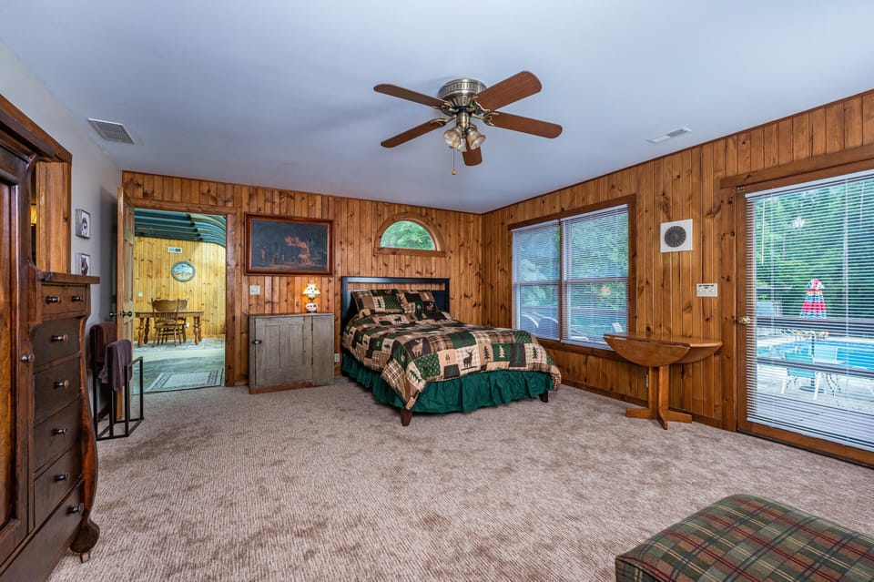 Spacious bedroom with wood panel walls, carpet flooring, ceiling fan, and sliding glass door leading to the pool area.