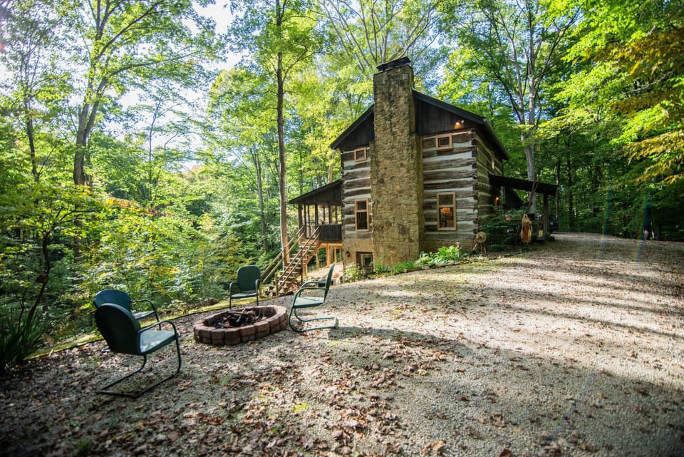 Outdoor fire pit encircled by four green metal chairs on a gravel patio beside the log cabin’s stone chimney.