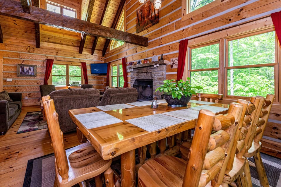 Dining area with a polished log table, matching chairs, and large windows framed by red curtains overlooking green woods.