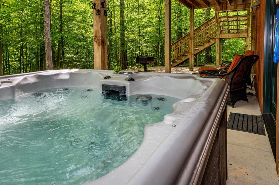 Bubbling hot tub on covered porch overlooks dense green forest, with wicker chairs and stairs nearby.