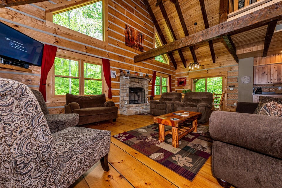Living room with wide plank wood floors, stone fireplace, patterned armchair, and large windows with red curtains.