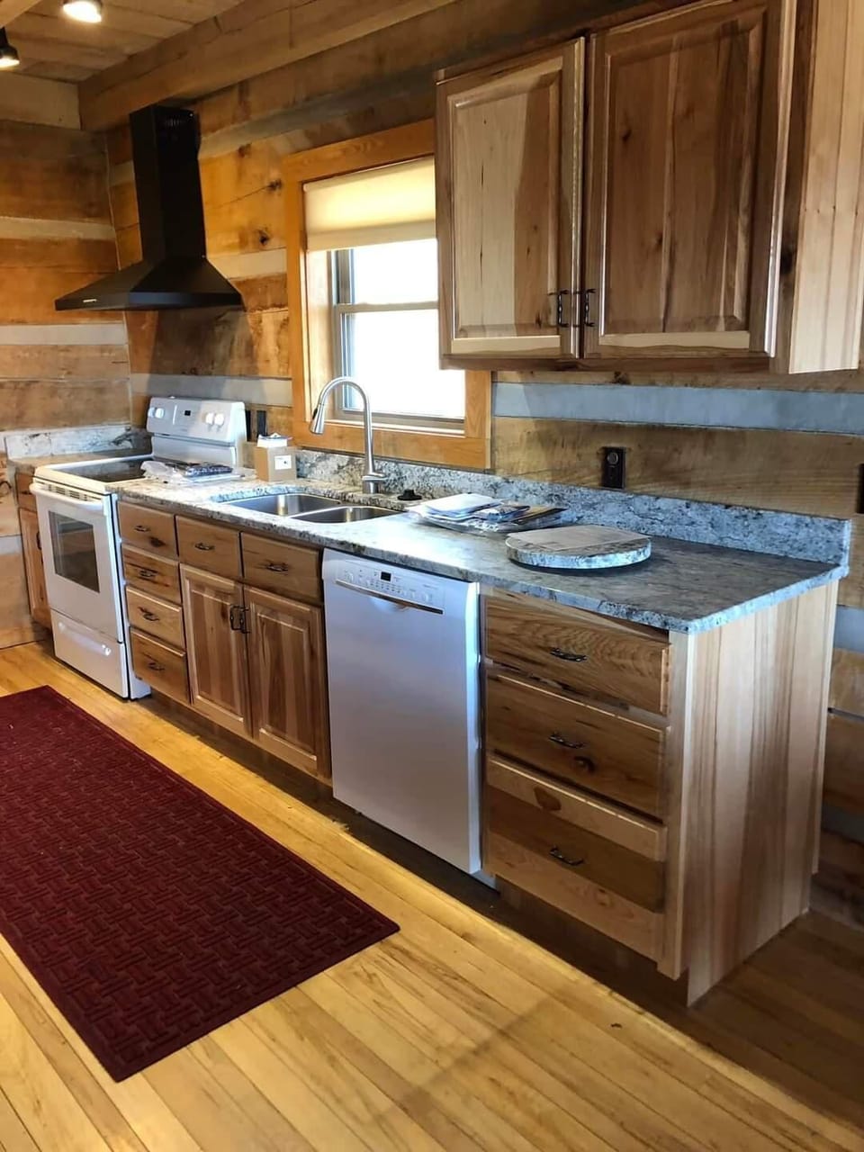 Natural wood cabinetry lines the kitchen with granite countertops, white appliances, and a red woven rug on hardwood floors.