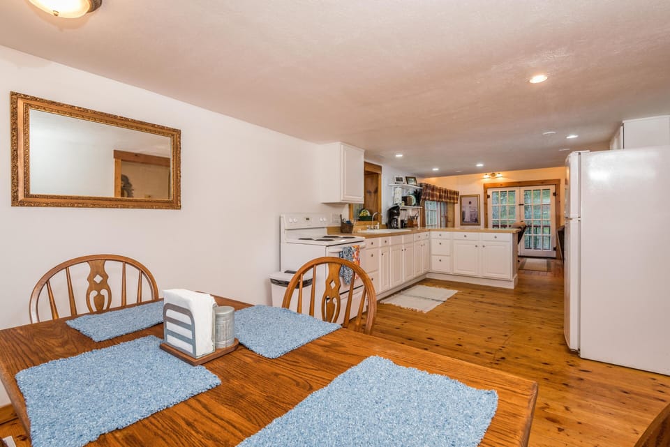 Dining table with blue placemats sits next to white cabinetry, wood floors, and a refrigerator in the kitchen area.