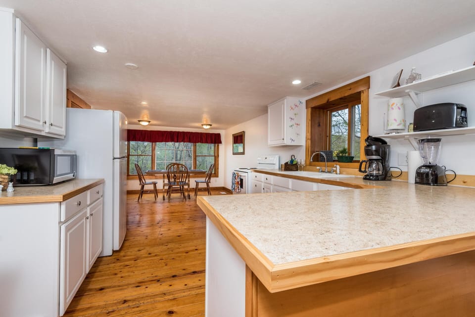 White cabinetry frames the kitchen with wood-trimmed countertops, a corner window, and a dining table by triple windows.