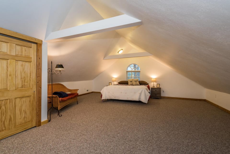 Attic bedroom under sloped ceilings with beige carpet, wooden closet door, wicker loveseat, and arched window.