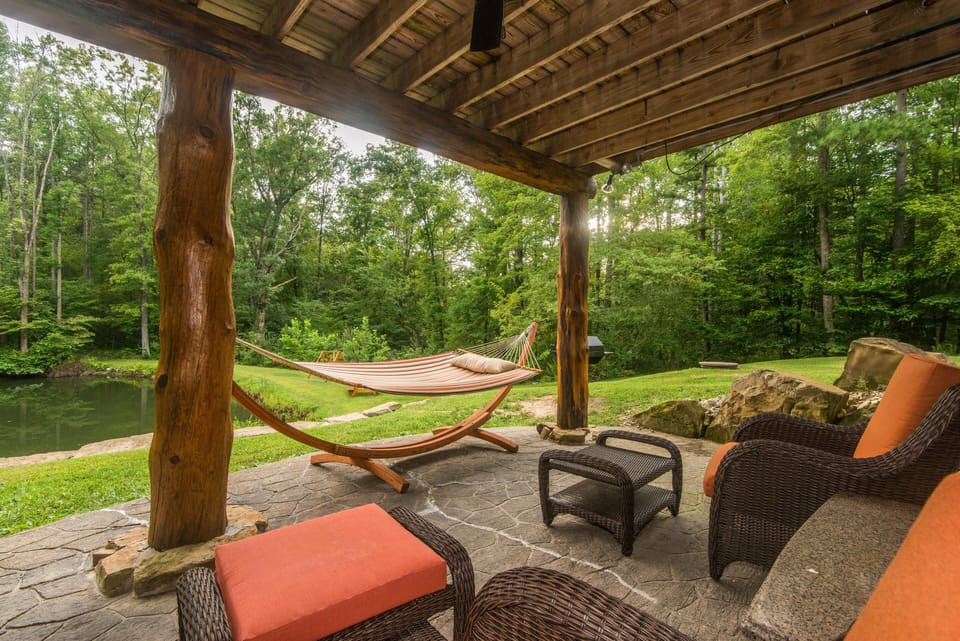 Covered patio with rustic log posts, wicker seating with orange cushions, and a striped hammock by the pond.