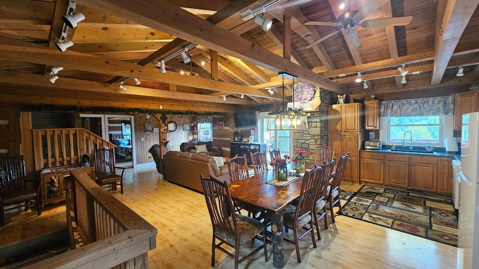Open dining area with a large rustic wood table and cushioned chairs beneath exposed wooden beams.
