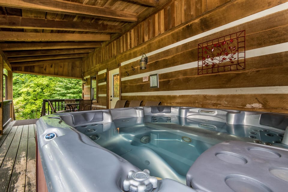 Covered porch with a large silver hot tub, wood plank flooring, log walls, and a small dining table overlooking trees.