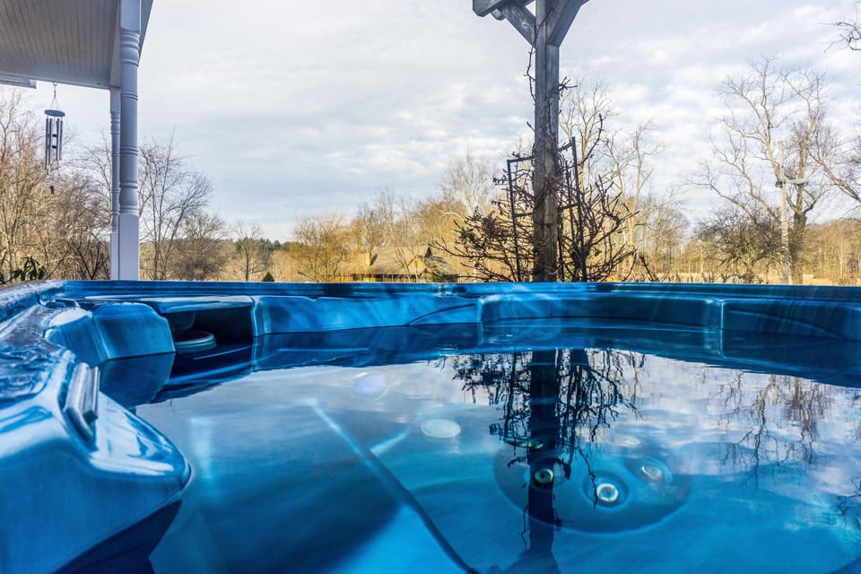 Blue hot tub under a covered porch reflects bare trees and a cloudy sky in the calm water.