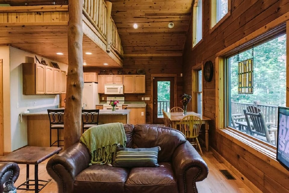 Leather loveseat with green throw faces large windows overlooking forested deck and dining nook.