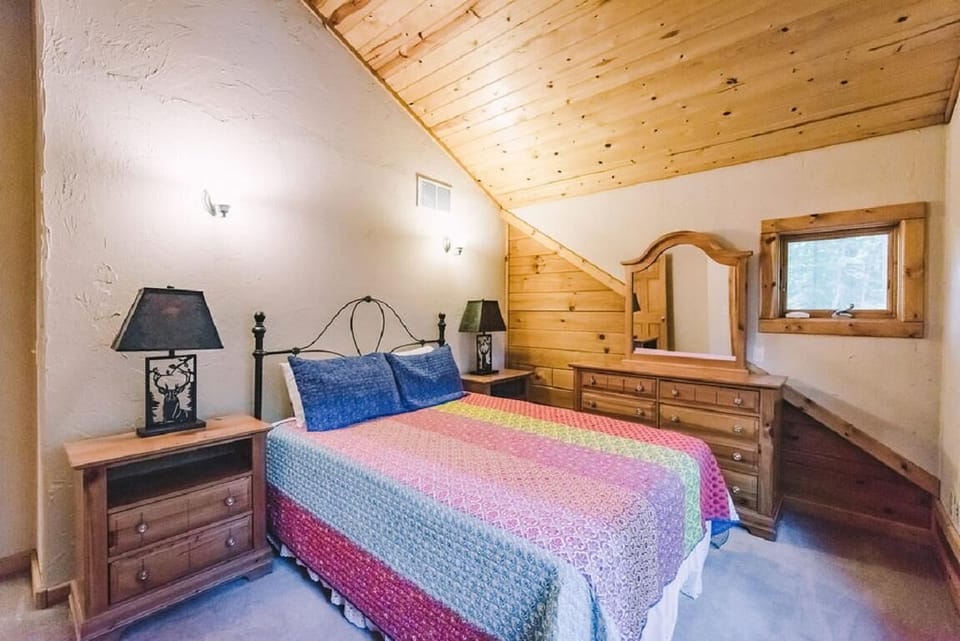 Iron bed with colorful quilt sits between wood nightstands topped with black lamps in sloped-ceiling bedroom.