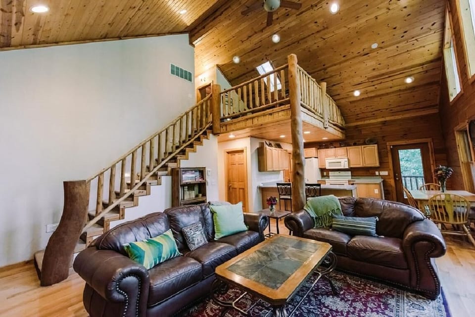 Dark leather sofas with colorful pillows face a slate-topped coffee table on a patterned rug beneath a vaulted pine ceiling.