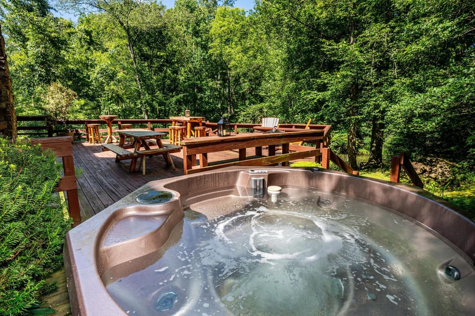 Bubbling hot tub sits on wooden deck surrounded by green trees, with picnic table and Adirondack chairs nearby.
