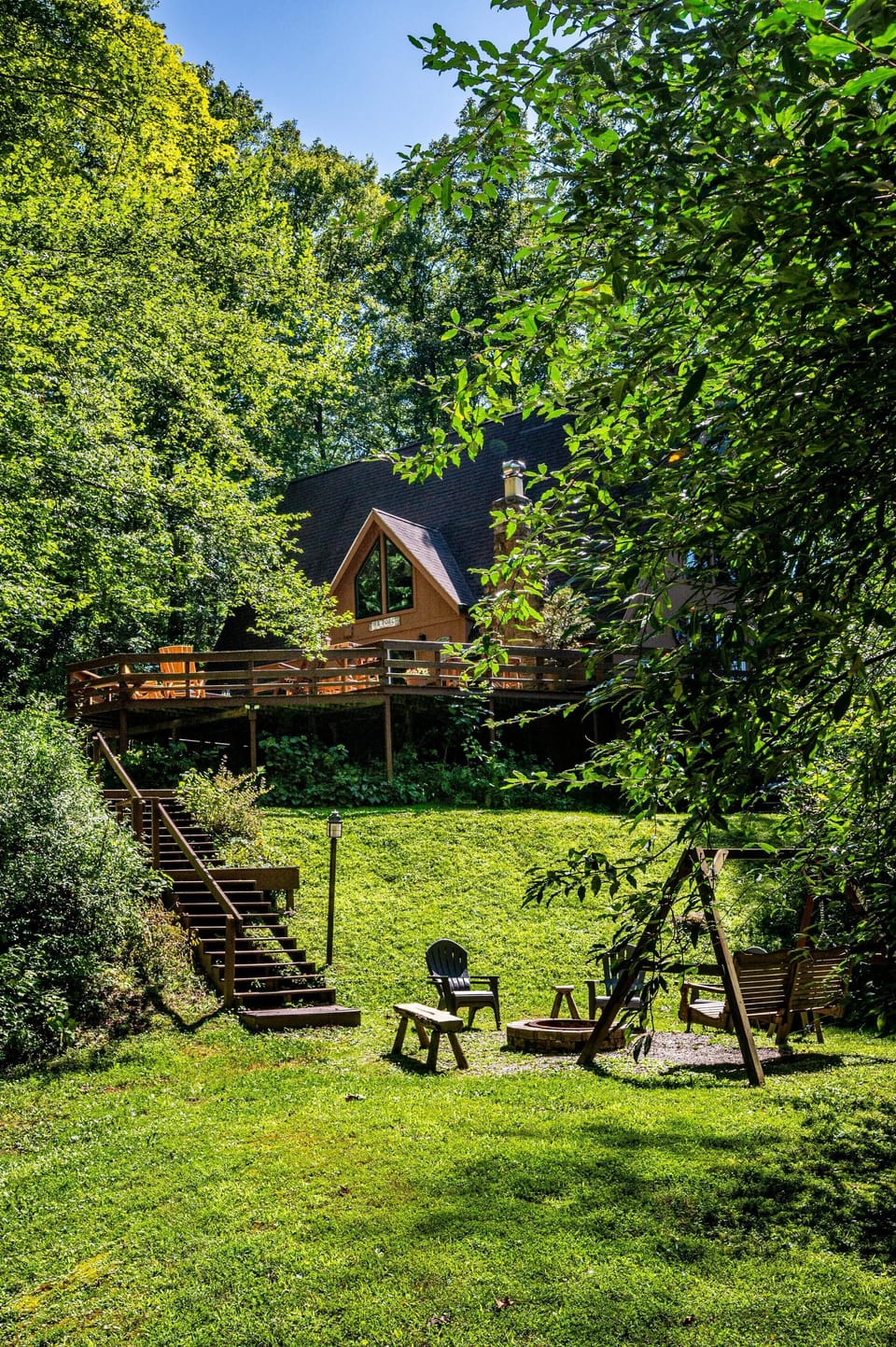 Wooden deck with Adirondack chairs overlooks grassy yard with fire pit, benches, and a wooden porch swing.