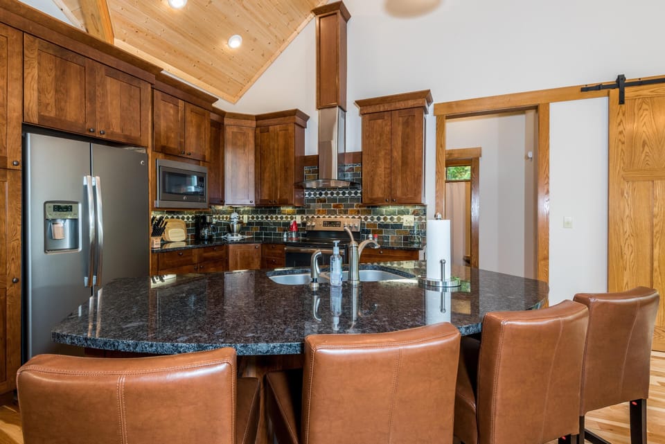 Dark granite countertop island with dual sinks and bronze faucets, surrounded by tan leather bar stools.