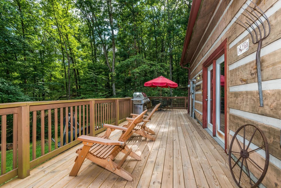 Spacious wood deck with two Adirondack chairs, stainless steel grill, red umbrella, and forest view.
