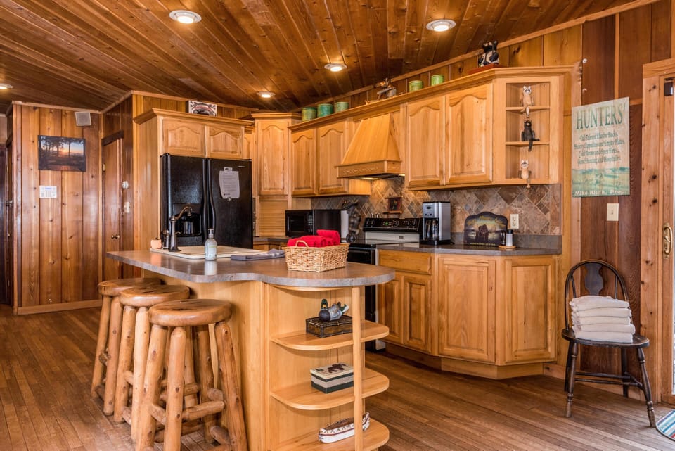 Kitchen with knotty pine cabinets, stone tile backsplash, black appliances, and a peninsula with log stools.