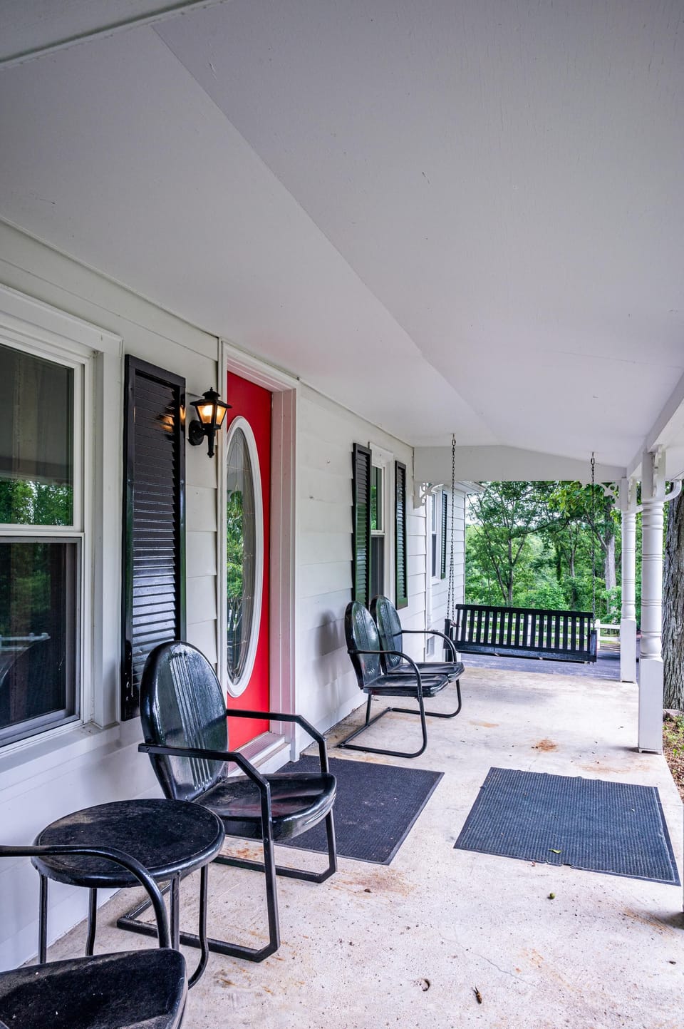 Front porch with black metal chairs, small round table, black porch swing, and bright red oval glass door.