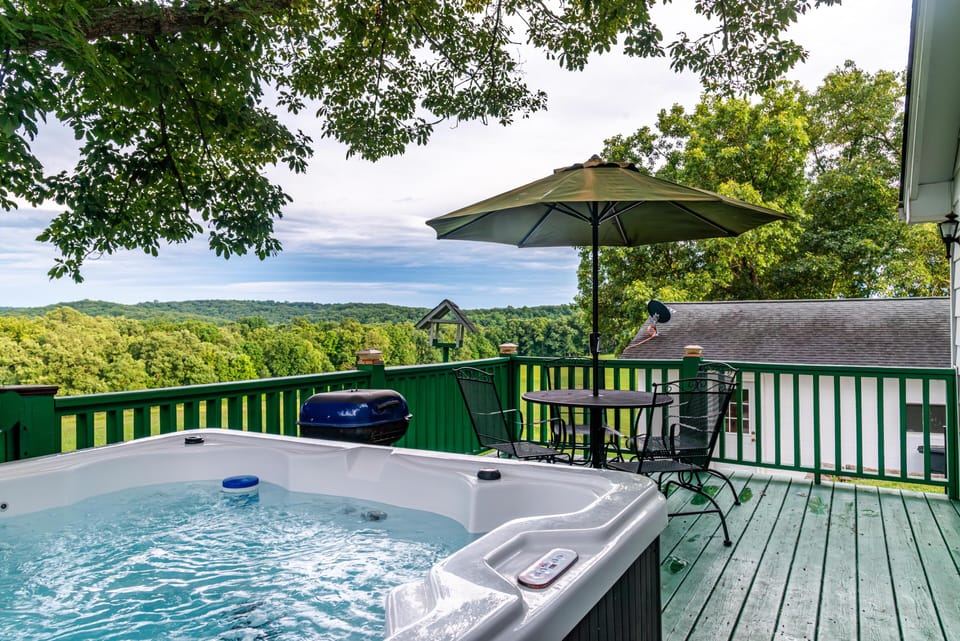 Hot tub bubbling beside a green wood deck railing, round metal table with umbrella, and forested hill views beyond.