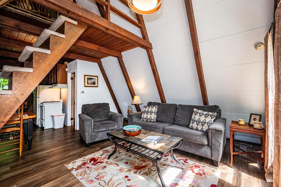 Living area with gray upholstered sofa and armchair, slate-tiled coffee table, and wood-paneled A-frame walls.