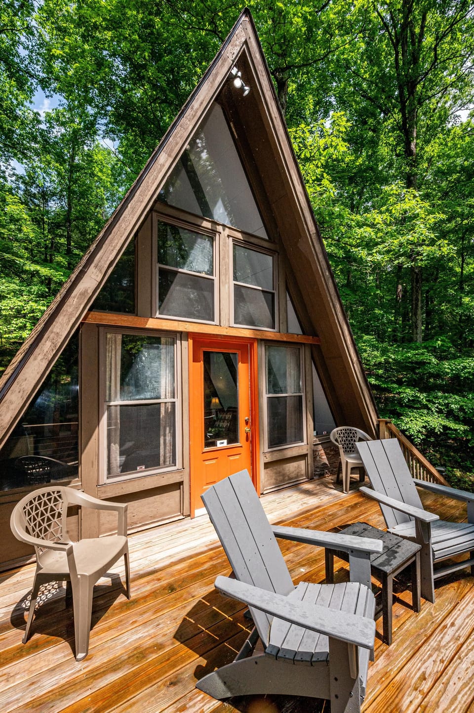Sunlit wooden deck with gray Adirondack chairs, beige plastic seats, and an orange door framed by large windows.